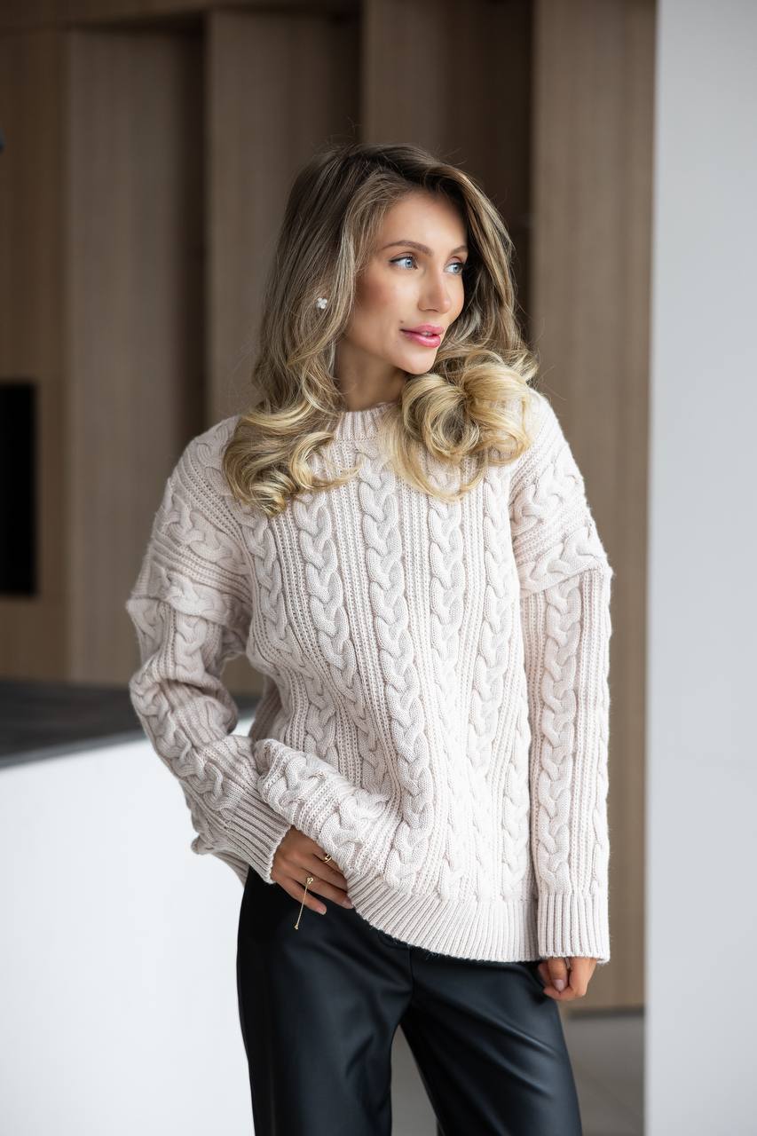 Woman wearing a cream cable knit sweater and black pants indoors.