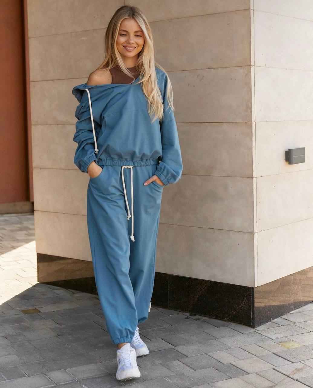 Woman wearing a blue tracksuit standing against a beige wall.
