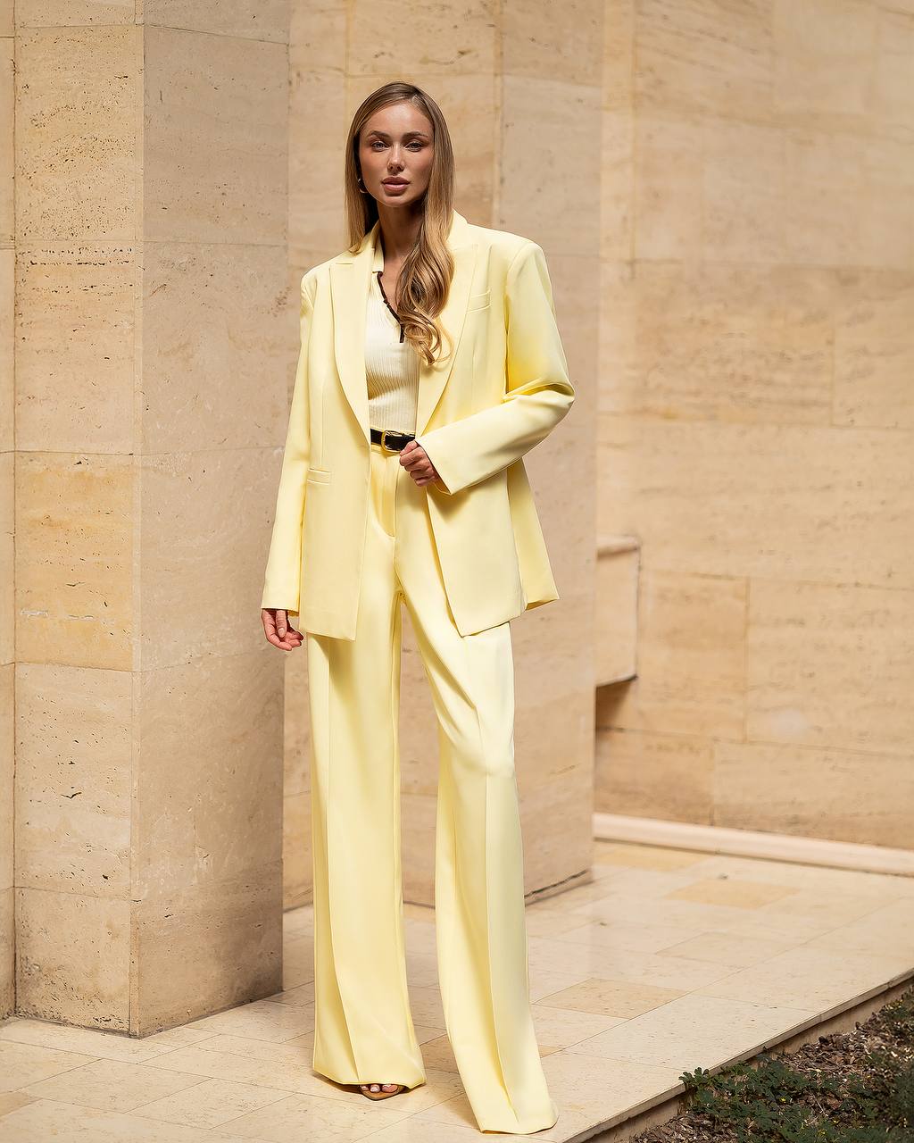 Woman in a light yellow suit standing against a beige stone wall.