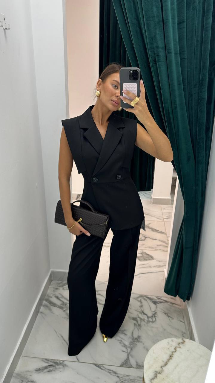 Woman taking a mirror selfie wearing a black sleeveless blazer and pants set, holding a black clutch.
