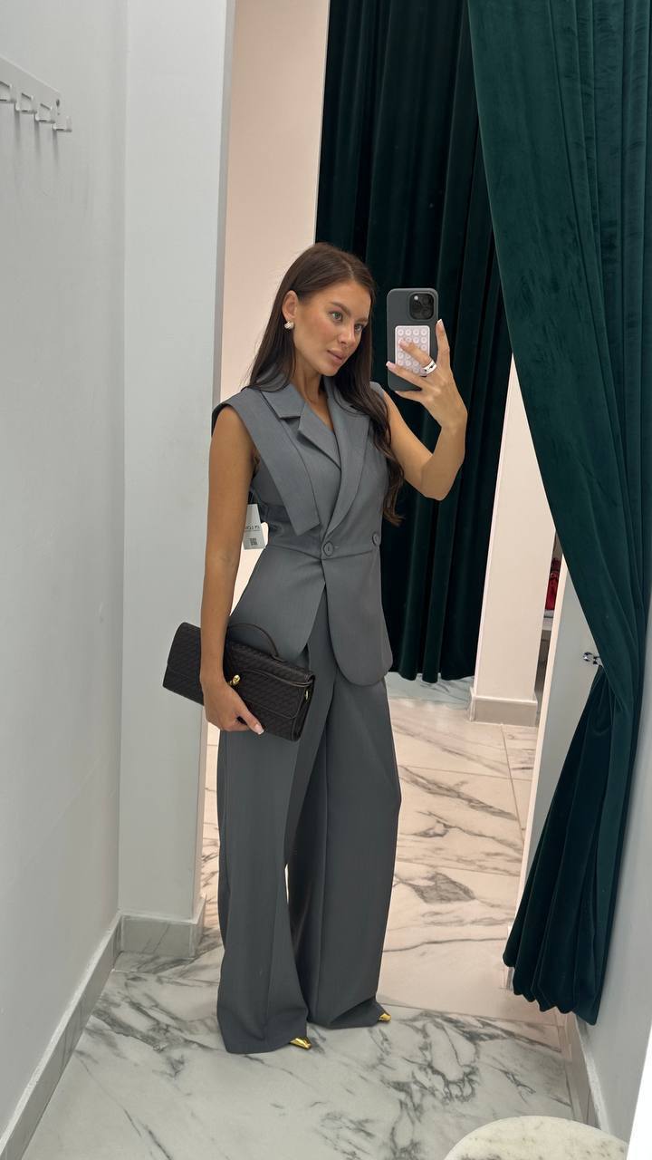 Woman taking a mirror selfie wearing a gray suit in a room with dark curtains.