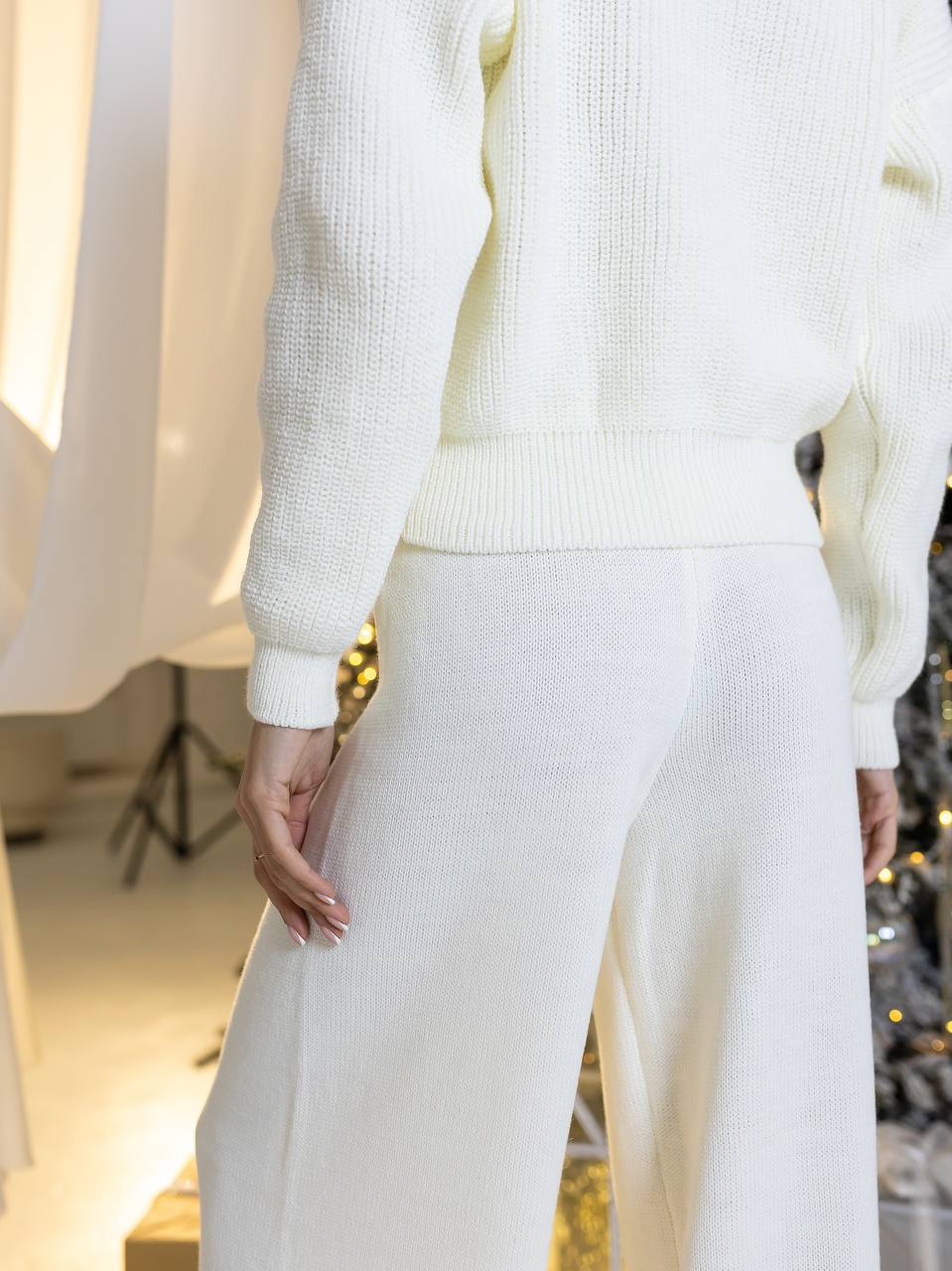 Person wearing a white knit sweater and pants in a softly lit room.