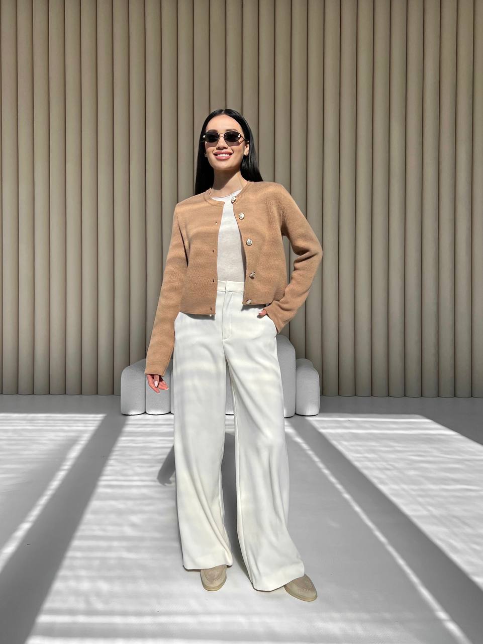 Woman wearing a brown jacket and white pants standing against a neutral background