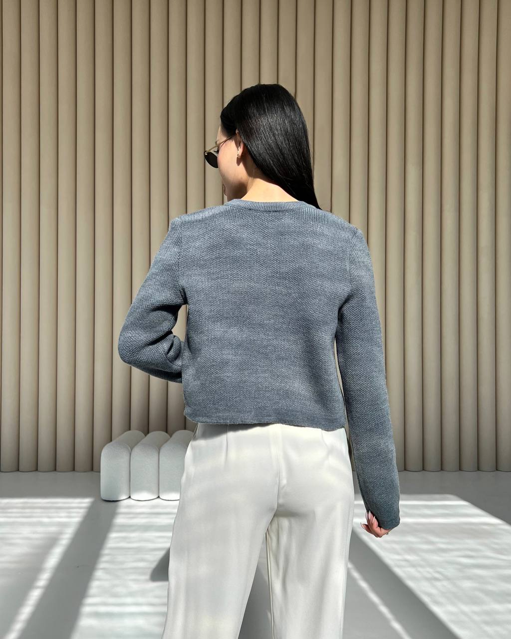 Woman wearing a gray sweater and white pants standing in a minimalistic room with beige walls.