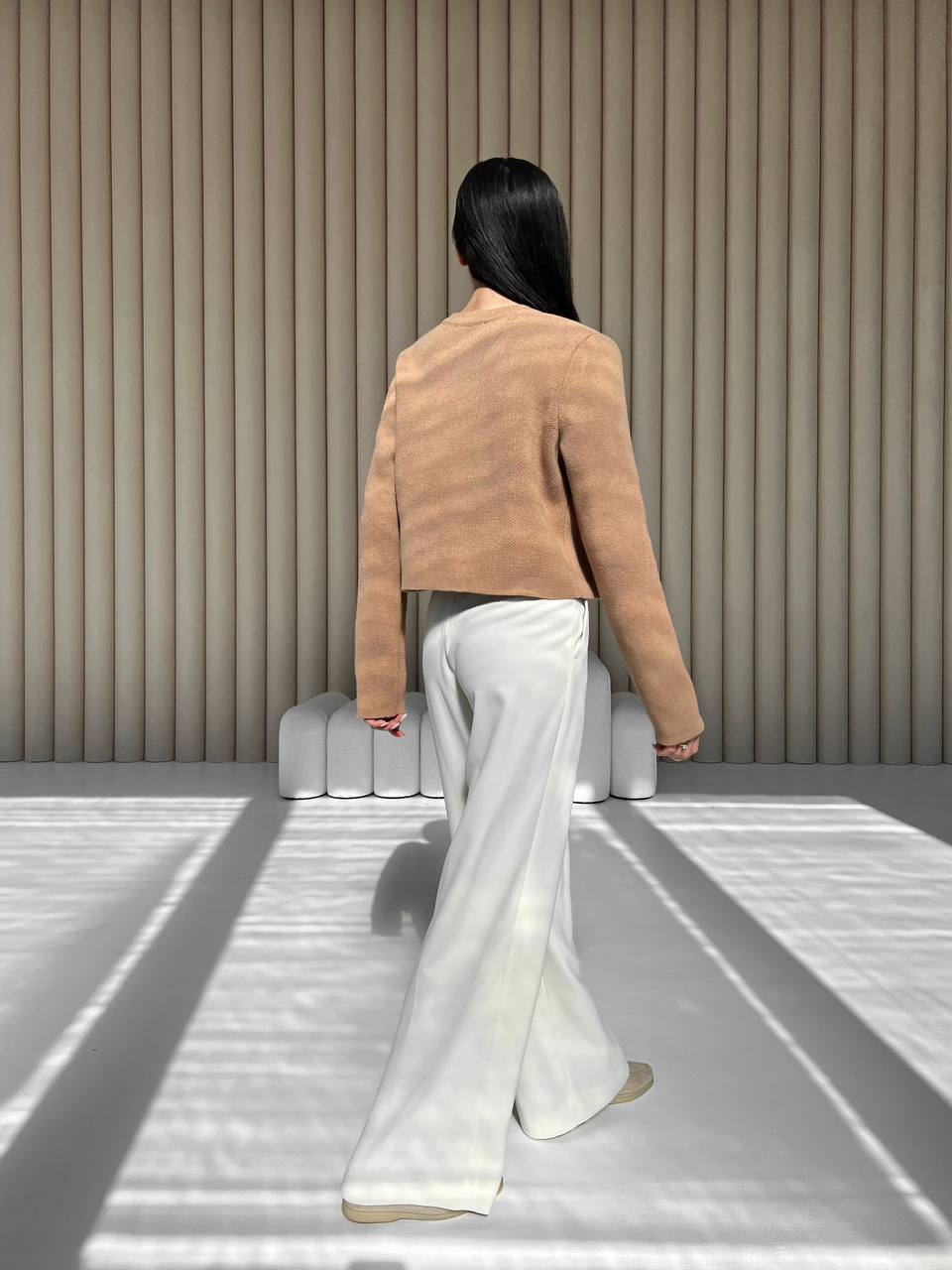 Person wearing a tan jacket and white pants standing in front of a textured wall.
