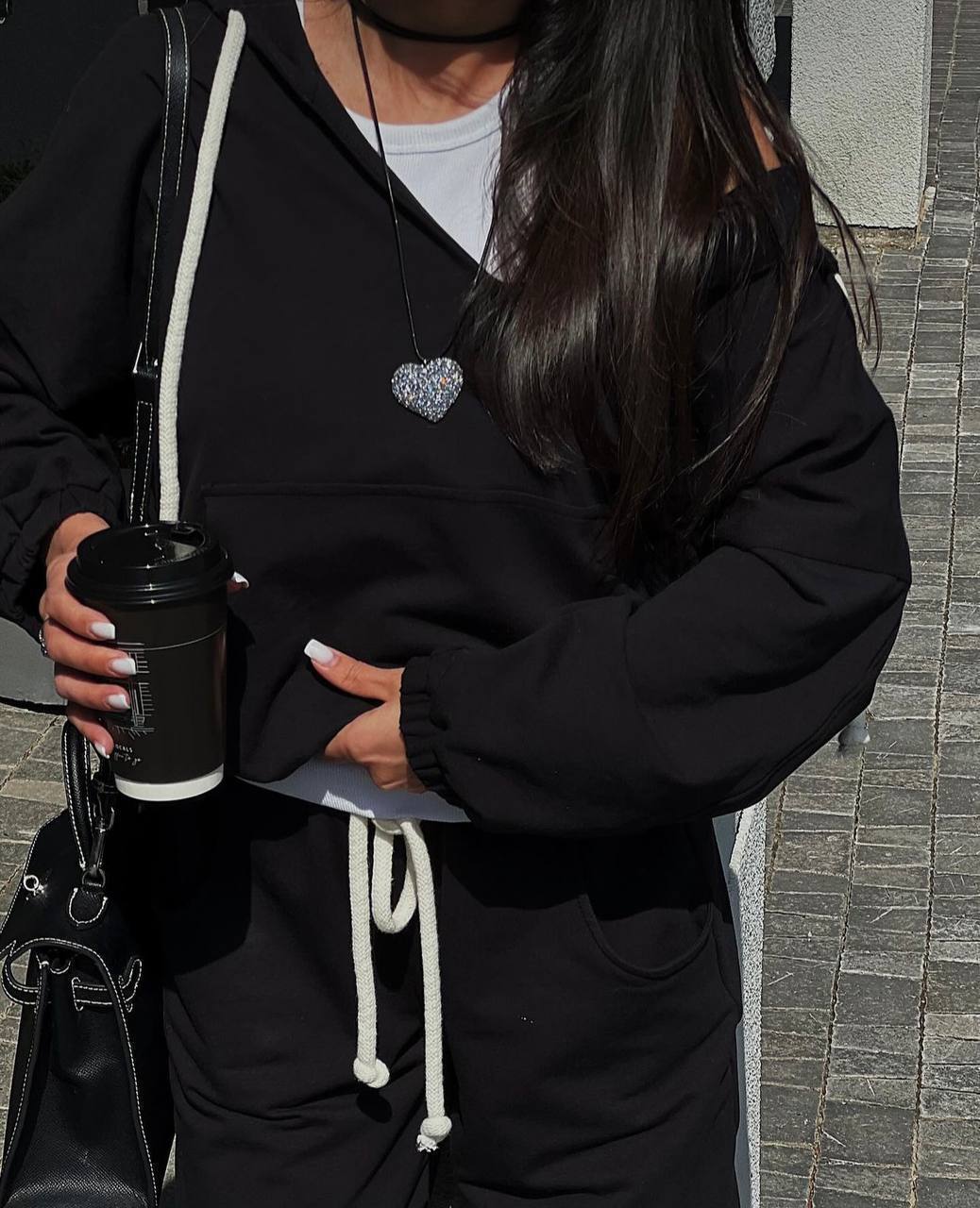 Person wearing a black hoodie with a heart-shaped brooch, holding a coffee cup.