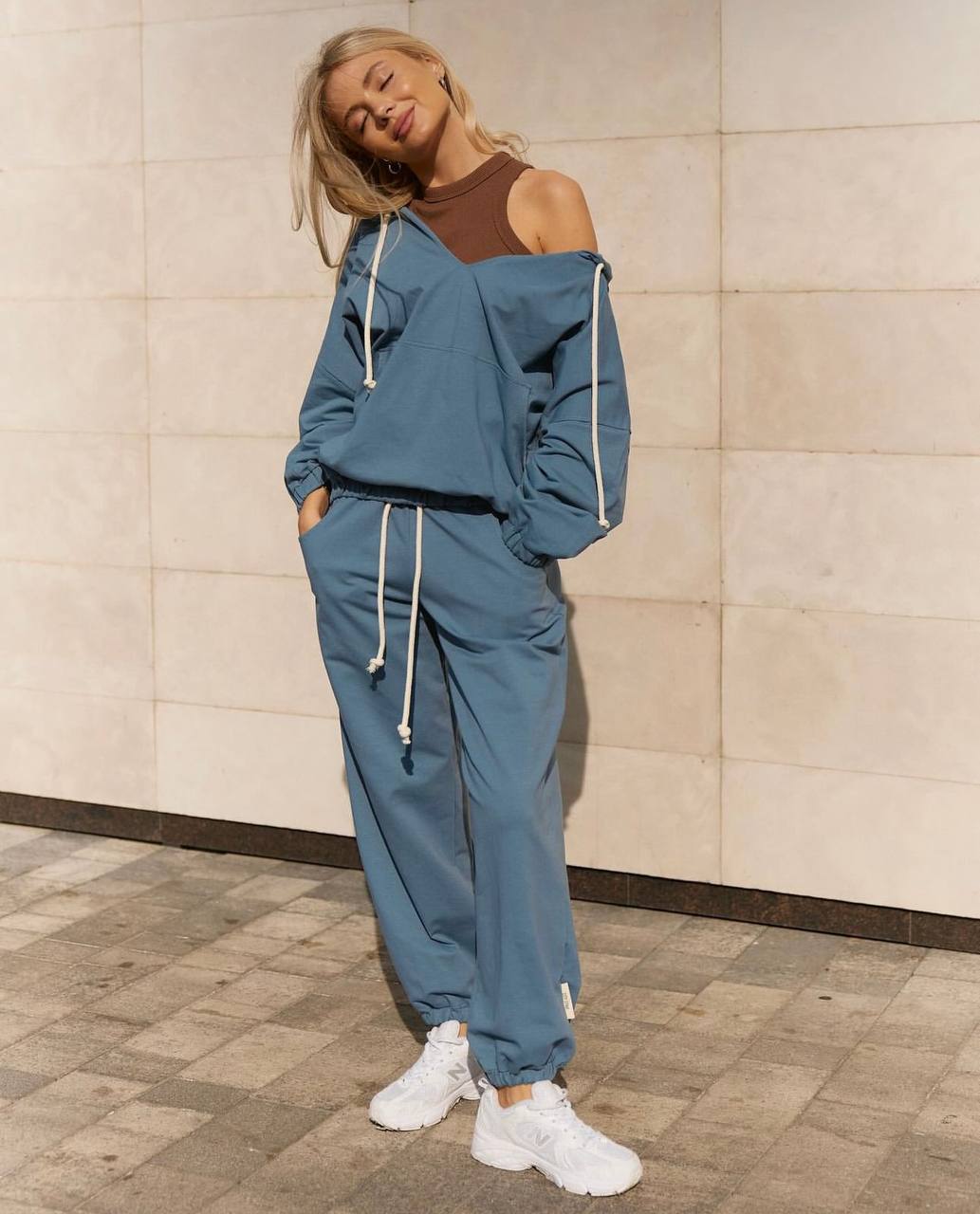 Woman wearing a blue tracksuit with white sneakers against a tiled wall.