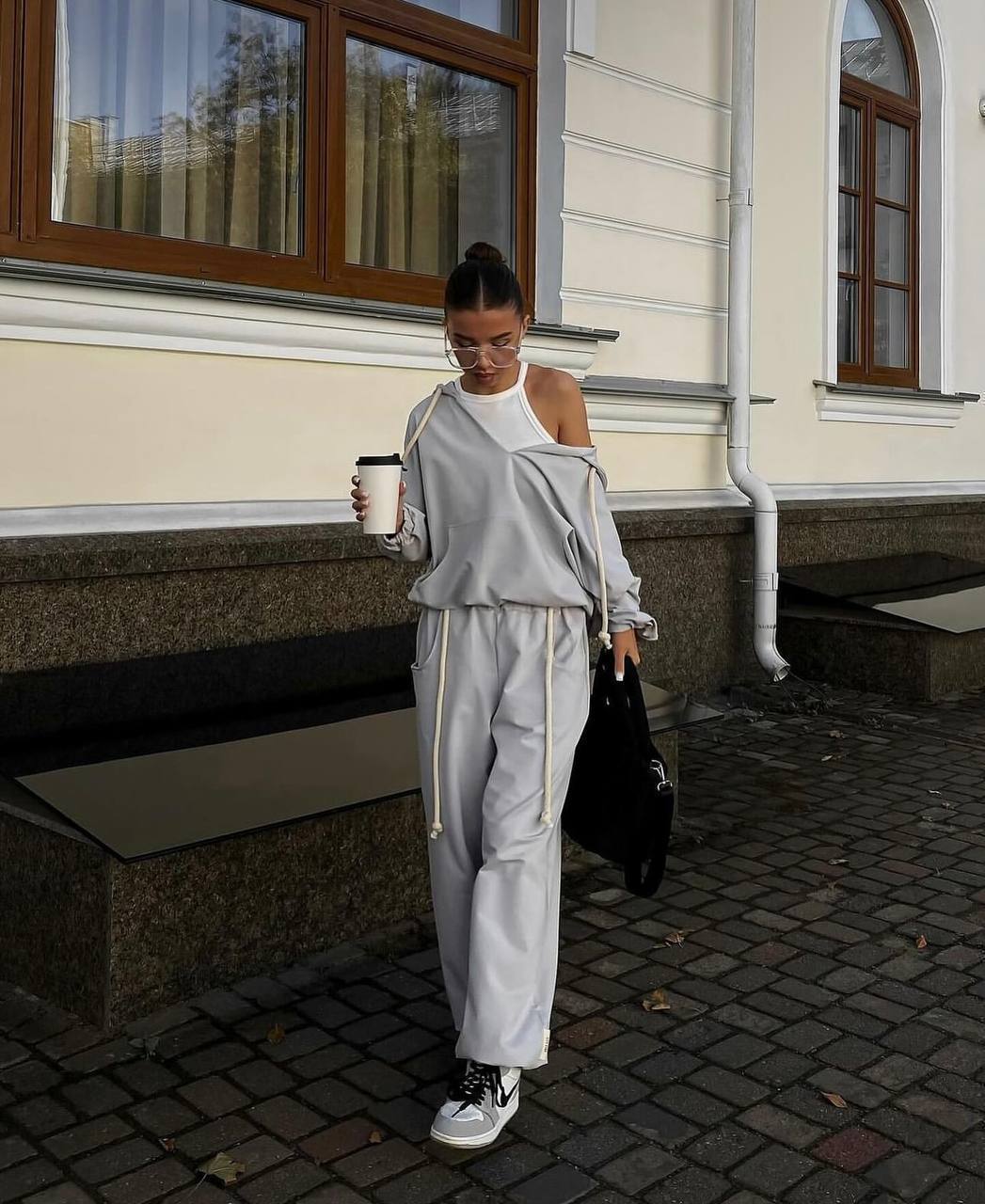 Woman in light gray outfit holding a coffee cup on a city street.